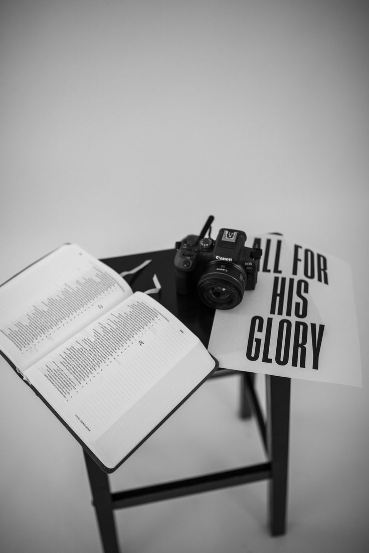 Black and white still life of an open Bible, a camera, and a poster reading All For His Glory
